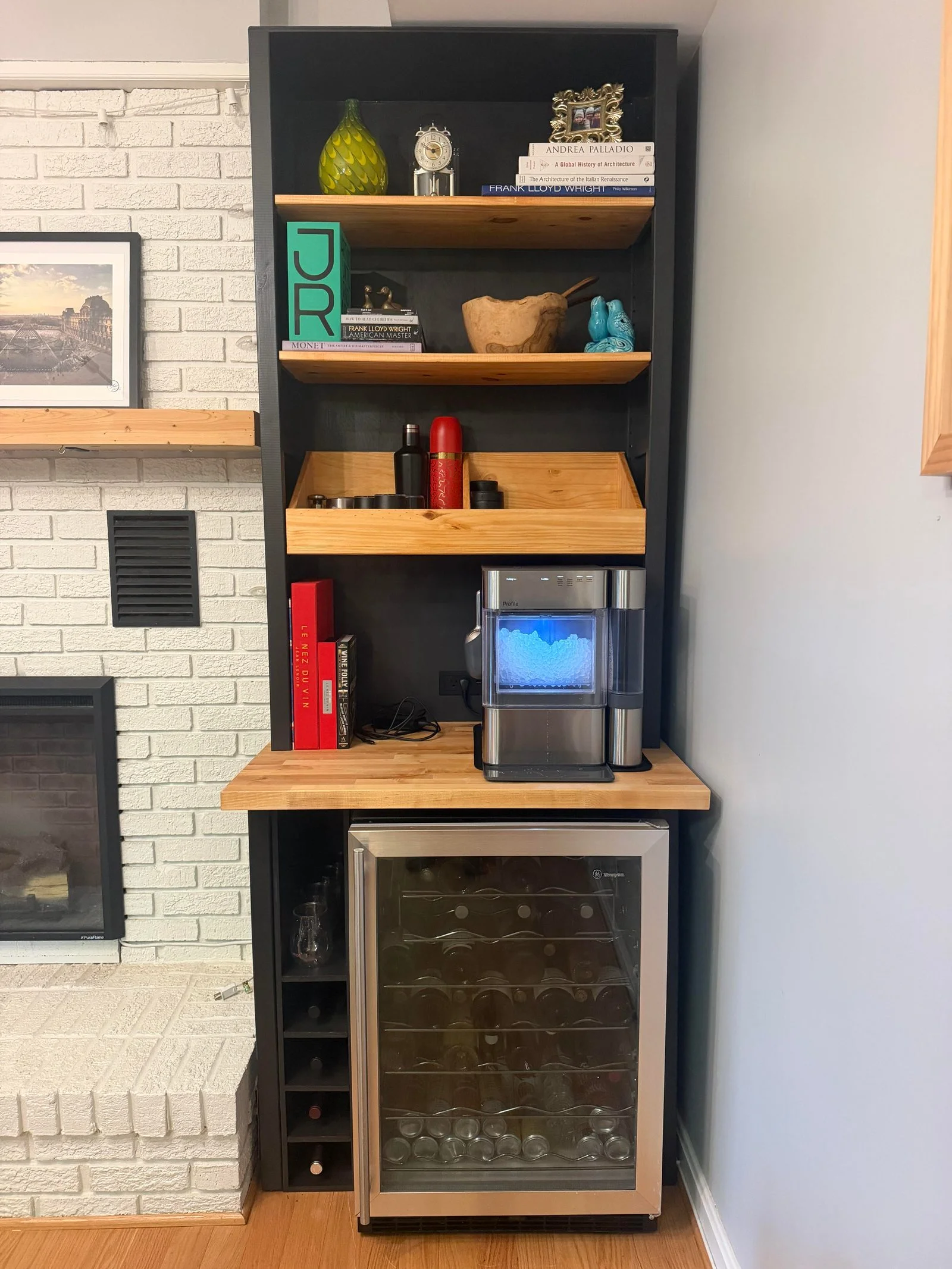 Libreria nera a incasso accanto a un camino in mattoni bianchi, con piano in legno e frigo cantina