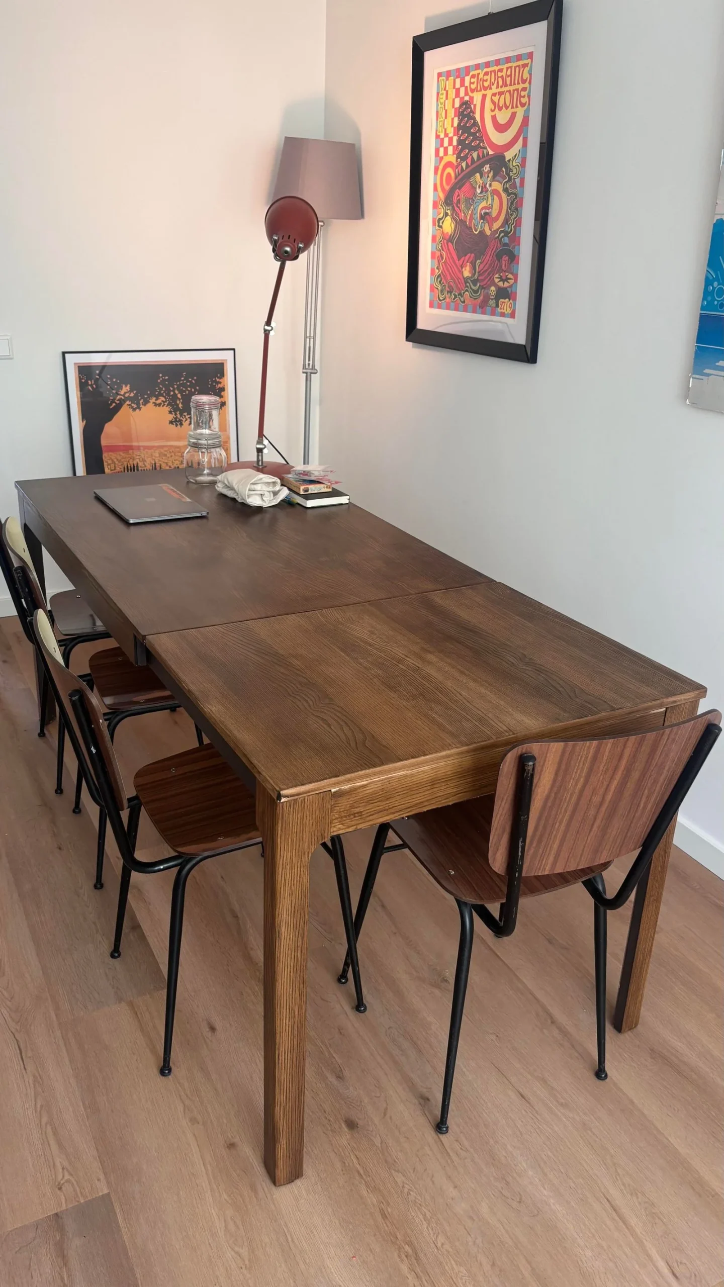 Tavolo da pranzo in legno color noce allungato, con sedie industriali, lampada e stampe incorniciate