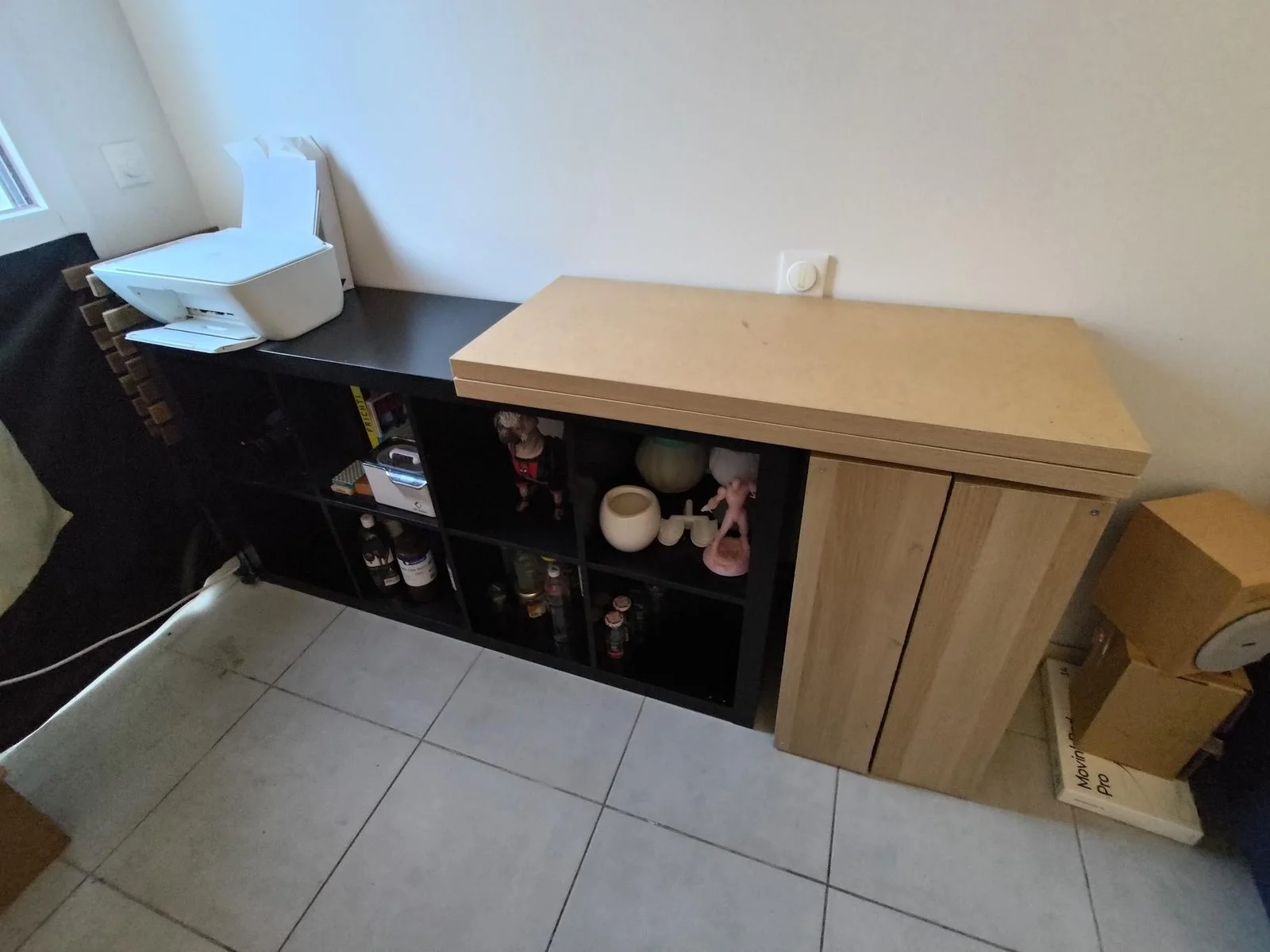 KALLAX bassa in rovere chiaro a muro con piano ripiegato sopra, accanto una KALLAX nera orizzontale