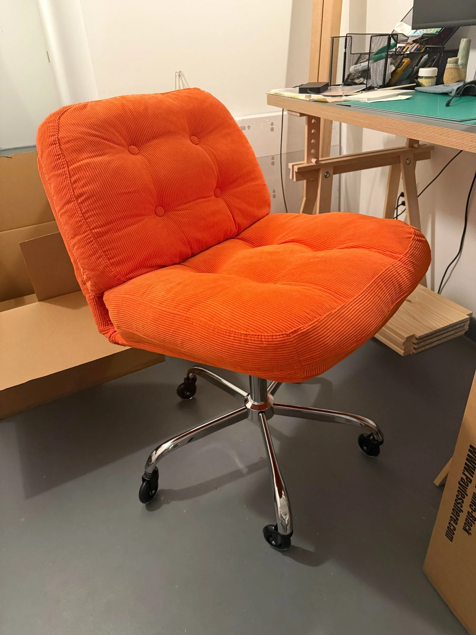 Poltroncina in velluto a coste arancione su crociera cromata con ruote accanto a una scrivania su cavalletti