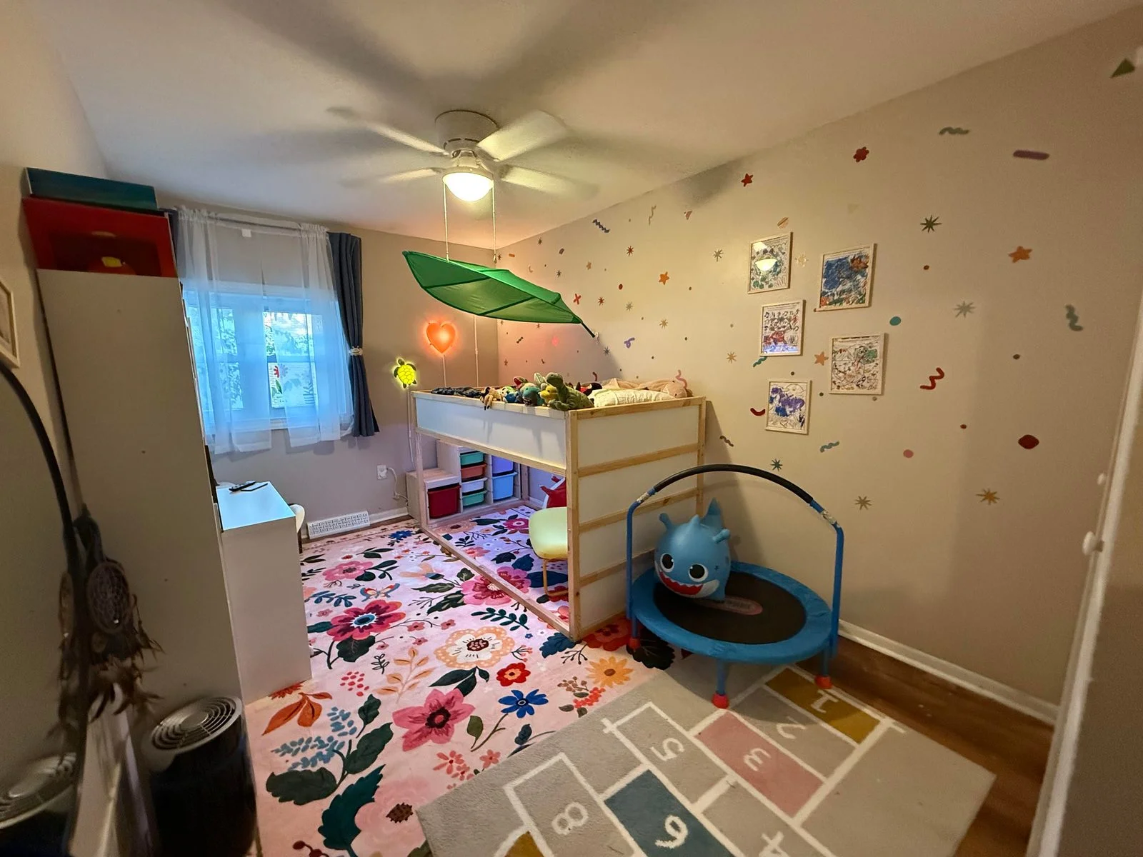 Vista d’insieme della cameretta con letto rialzato, contenitori sotto il letto, finestra e tappeto a fiori